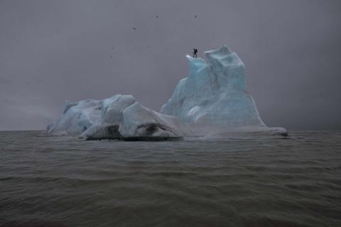 An artwork depicting a blue-grey iceberg against a dark grey sea and sky; a person stands on top of the iceberg, melting its tip with a blowtorch.