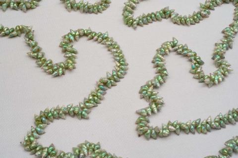 A photograph of a necklace made from small, shiny green shells, arranged in river-like wiggles over a white surface.