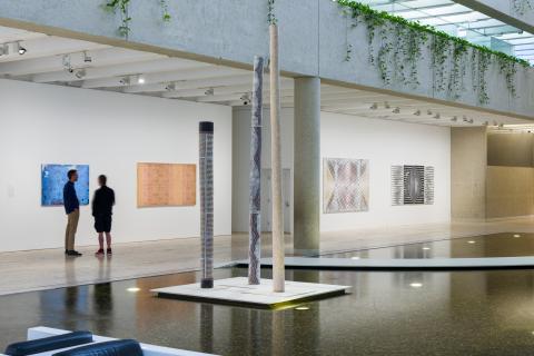 An installation view of a gallery space set over an indoor pond or water feature, on which three memorial poles are displayed on a plinth.