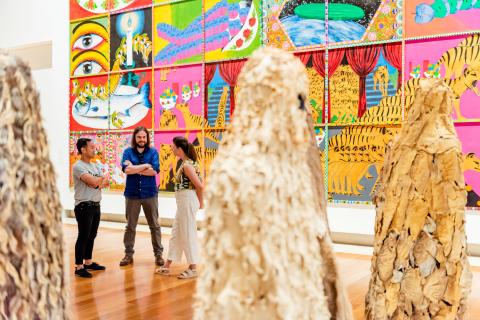An installation view of a gallery space featuring a very large-scale, colourful work on the wall and, in the foreground, human-like sculptures made from frog and fish skins. Some visitors discuss the work in the background.