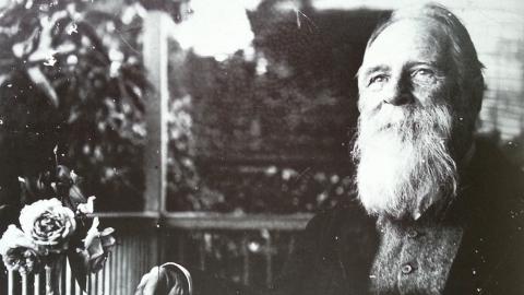 A black-and-white photograph of an older man seated on a verandah in the late 1890s; he is smiling gently, and seated with his hand at an open book and with a small vase of roses at the table.