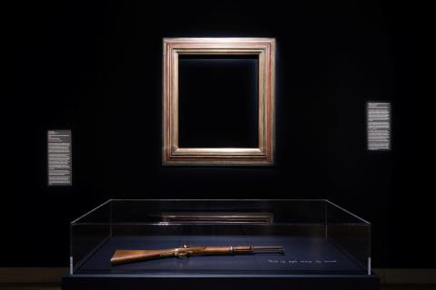 An installation view in a dark room with gallery walls painted black; an empty gold frame on the wall, in the centre of the photo, frames the darkness; beneath it, a rifle is displayed in a glass cabinet.