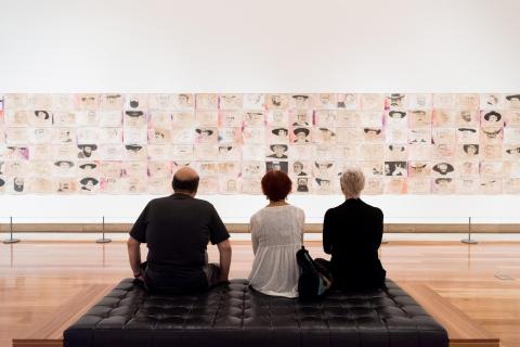 An installation view of a large work composed of many smaller drawings and collages on a white gallery wall, with three seated visitors looking on.
