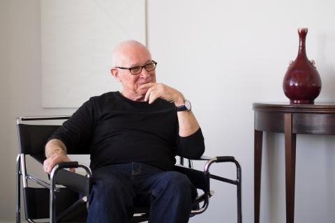A photograph of a bald man in glasses, a black sweater and dark jeans sitting and looking away, as if in thought.