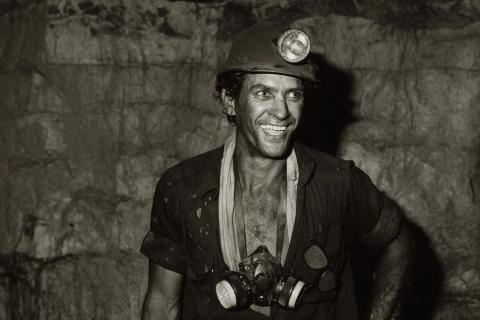 A black-and-white photograph of a miner, who is grinning and wearing a hat with a lamp on it. A mask is slung around his neck.