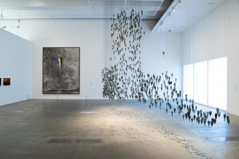 An installation view of a gallery space, with a glass sculpture suspended from the ceiling at right, by a large window.