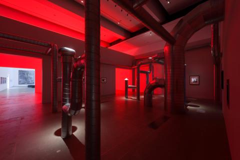 An installation view of a red-lit gallery space, with an artwork composed of piping criss-crossing the space.