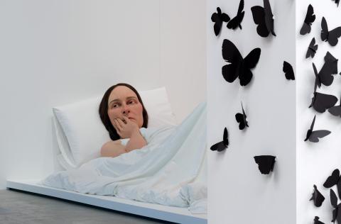 An installation view of a realistic sculpture of a woman lying in bed, with a wall covered in black butterflies at right.