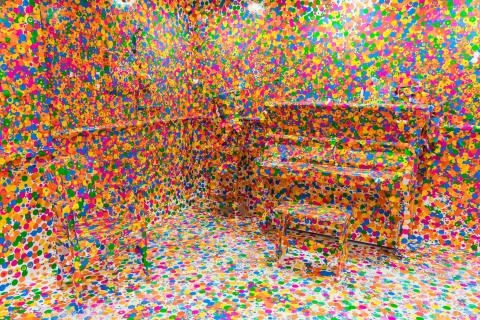 An installation view of a room with a piano, but every surface is covered up with colourful dots.