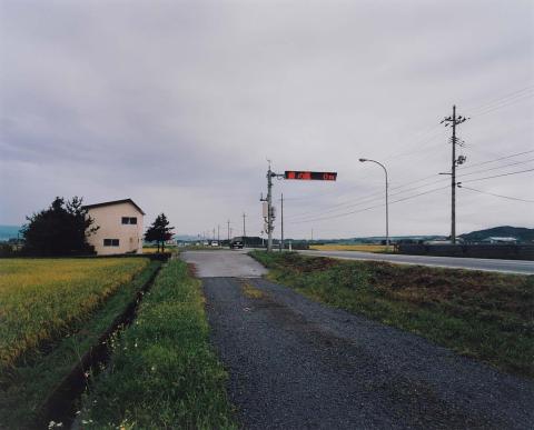 A photograph of a rural road in Japan, with a house seen at left and fields either side.