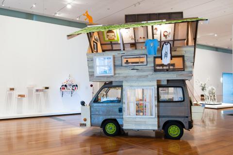 An installation view of a bright gallery space, with a sculptural artwork taking the form of a van carrying a cobbled-on second level on top.