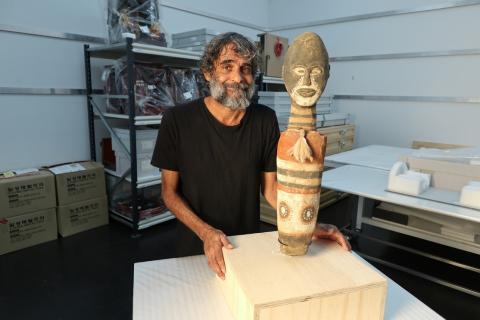An Australian Indigenous man stands with a sculpture, which depicts a small male figure, around 60cm tall; the photograph is taken in a room with white walls, possibly an art conservation space.