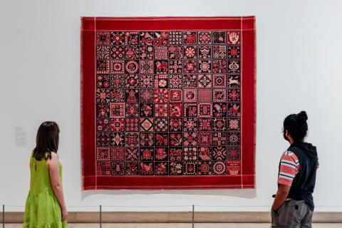 An installation view of a textile work in red tones on a white gallery wall, with two visitors looking on.