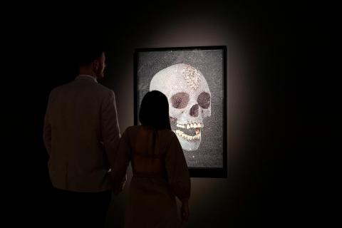 Installation view of a dark gallery space, with two visitors viewing an artwork depicting a sparkling skull.