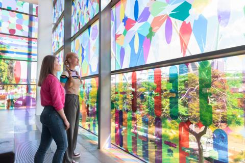 Two people stand looking at and through Shannon Novak's transparent vinyl window installation, with many colours casting rainbows on the floor of the river room.