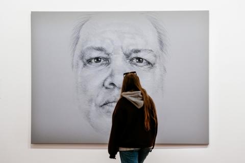 An installation view of a large charcoal drawing of a face on a white-grey background, with a visitor looking on.