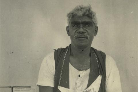 A black-and-white photograph of an Australian Indigenous man wearing a boomerang-shaped 'king plate' around his neck, along with a t-shirt and vest