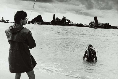 A black-and-white photo of a man fishing, with a view of a scuba diver and a shipwreck in the distance