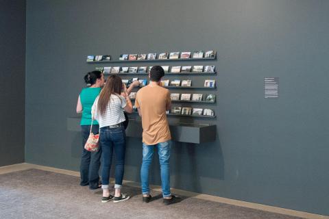 An installation view of a work comprised of postcards displayed on shelves on a wall, with three visitors looking on up close.