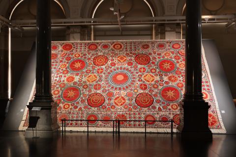 Madina Kasimbaeva / Uzbekistan, b.1981 / Palak suzani 2016–21, installed at Art Museum RIGA BOURSE, Latvia, 2024 / © Madina Kasimbaeva / Photograph: Oksana Mizgajeva / Image courtesy: The Latvian National Museum of Art and Art Museum RIGA BOURSE A large, red-patterned textile work installed in a gallery space with grand architectural pillars