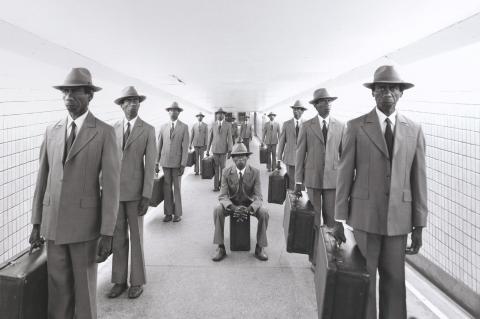 A detail view of a greyscale photograph in which the same figure, dressed in a vintage suit, repeats multiple times in different commuter-coded poses inside a pedestrian tunnel