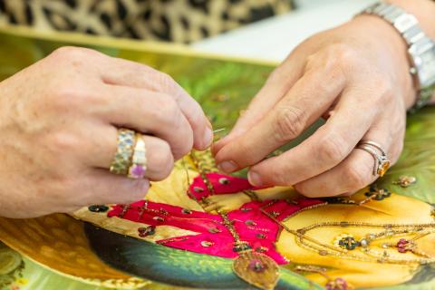 A photograph documenting conservation work on an embroidered oleograph by Raja Ravi Varma, showing just the textile conservator's hands carefully stabilising embroidered elements of a colourful print.