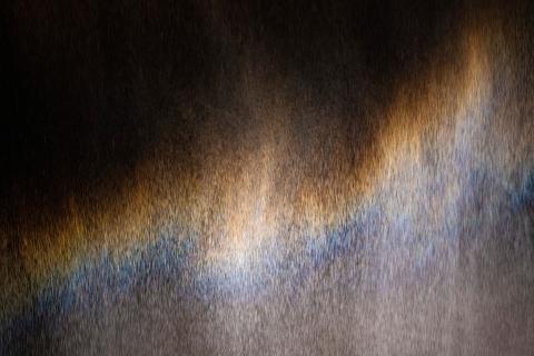 An artwork comprised of a vertical sheet of fine mist illuminated with soft blue, amber and violet tones beneath a small overhead light.