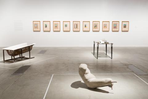 An installation view of sculptural works (foreground) and miniature paintings (background) installed in a bright gallery space