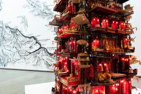 A sculptural work resembling a tall shrine, with a view of a map-like mural in the background, installed in a bright gallery space