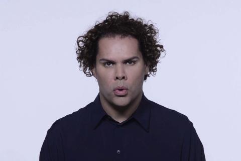 A still drawn from a video work, in which we see the artist Dr Christian Thompson – an artist of Bidjara/Irish/Chinese–Australian heritage – standing in front of a bright background. He appears to be in the middle of singing. He has short, curly hair and wears a dark blue button-up shirt.