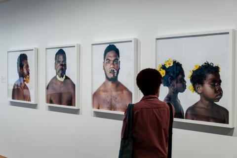 A photograph of a visitor walking past an installation of photographs on a white gallery wall; the photographs depict portraits of Indigenous Australian men adorned with flowers.