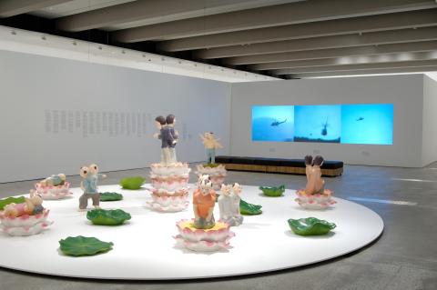 A view of works installed in a bright gallery space, including a series of sculptures installed on a circular plinth, with video works installed on the back wall