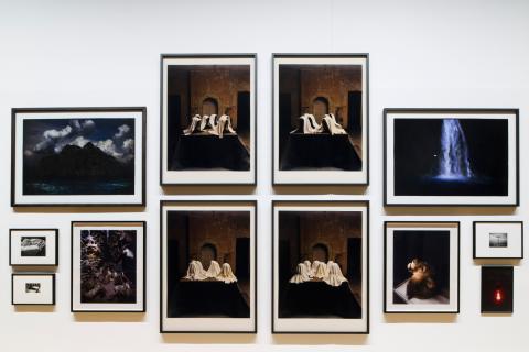 This photograph captures a view of a salon-style installation of photographs, with very small works hung at the far edges and the largest works hung in the middle. Multiple artists are represented; however, each photograph included has dramatic, Baroque elements in its composition.