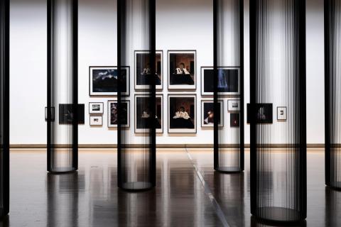 This photograph captures a view of a salon-style installation of photographs, with very small works hung at the far edges and the largest works hung in the middle. In the foreground, columns made of tape from inside VHS cassettes appear both monumental and transparent. Multiple artists are represented; however, each photograph included has dramatic, Baroque elements in its composition.