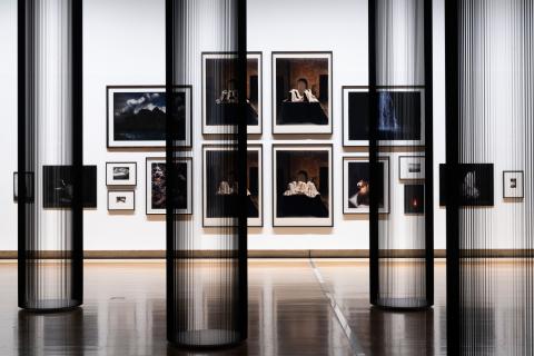 This photograph captures a view of a salon-style installation of photographs, with very small works hung at the far edges and the largest works hung in the middle. In the foreground, columns made of tape from inside VHS cassettes appear both monumental and transparent. Multiple artists are represented; however, each photograph included has dramatic, Baroque elements in its composition.