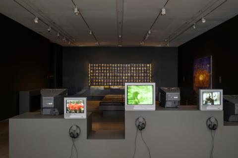 An installation view of a dark gallery space, with video works shown on CRT televisions in the foreground, and with glimpses of works installed on the walls in the dimly lit gallery behind.