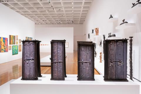 A view of an installation of small, black-coloured wooden doors, with a bright, white-walled gallery behind