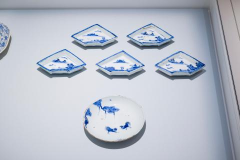 A photograph of kosometsuke works, made in China during the Ming Dynasty for the Japanese market: a set of five small blue-and-white dishes are installed in a vitrine above a larger, round dish decorated with fighting cockerels. They are installed on a very light blue background within the vitrine.