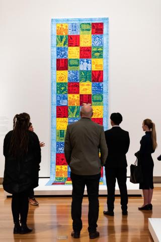 A photograph of a brightly coloured, portrait-oriented textile installed in a gallery space; four gallery visitors look on