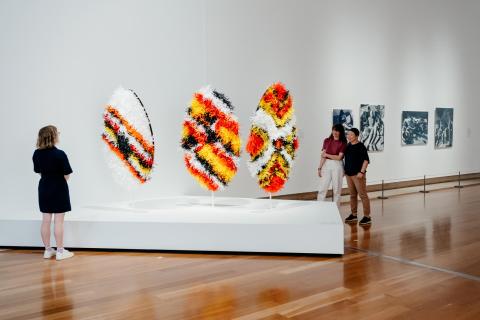 A view of three, large shield-like sculptures made from orange, black and yellow materials, installed in a bright gallery space; gallery visitors look on from either side of the installation