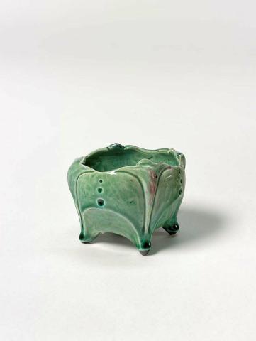 Artwork Footed bowl this artwork made of Hand-built earthenware with in-curving sides and triform shapes extending to form feet.  Three graduated dots set above a carved shape on each side.  Glazed green with red, created in 1920-01-01