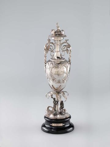 Artwork Racing trophy:  The Wythes and Hodgson Cup this artwork made of Narrow necked ovoid body arising from a tree fern stem with incurving handles and cast grapes, parrots and snake decoration.  The lid surmounted by a kangaroo.  The beaded heptafoil base with repousse rocks, ferns and figures of a male and female Aborigine in oxidised silver.  Engraved on one face with an inscription between repousse fern trees and on the other with three horses in full gallop.  Mounted on a black turned wooden base with metal plaque, created in 1870-01-01