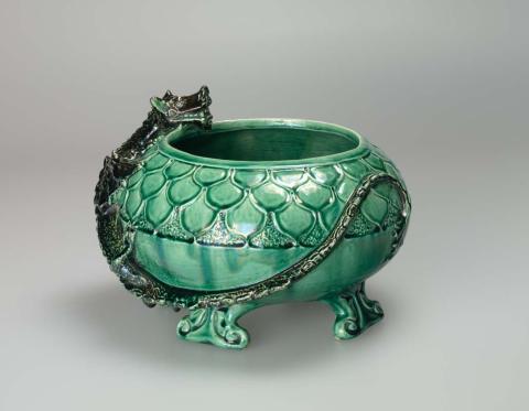Artwork Dragon bowl this artwork made of Earthenware, hand-built, flattened spherical bowl set on four scrolled feet. The rim decorated with a carved scale pattern and a modelled dragon with head overlapping the rim and extending around the body. Glazed green with the dragon in black, created in 1940-01-01