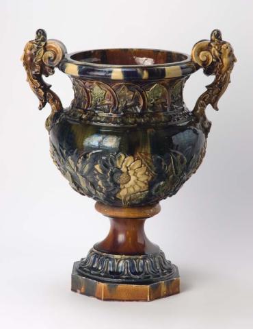 Artwork Jardiniere this artwork made of Earthenware white clay body.  The neck modelled with ivy leaves within arches, the swelling body with four sunflowers and leafy branches and the handles modelled as two grotesque masks.  Glaze splashed clear brown, green and deep cobalt
