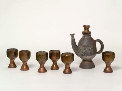 Artwork Wine jug and six goblets this artwork made of Earthenware, thrown. The jug with erect flaring spout and semicircular handle on waisted base. Glazed mottled manganese green with pewter base and bronze handle. Goblets glazed mottled manganese with bronze foot