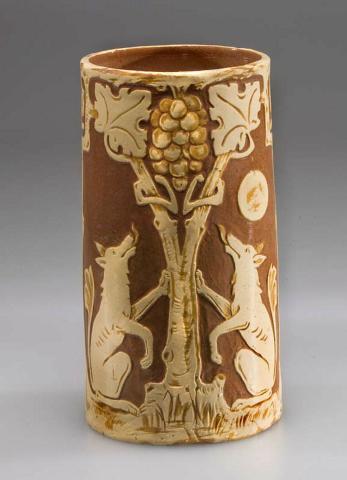 Artwork Cylindrical vase:  (The fox and the grapes) this artwork made of Hand-built brown clay cylindrical shape, dipped ochre and cream clays and carved with a formalised design.  Clear glaze, created in 1933-01-01
