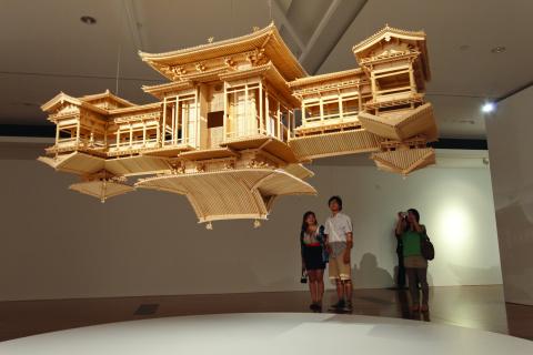 An installation view of a sculpture in the form of a bamboo temple suspended from the ceiling in a gallery space, with onlookers behind.