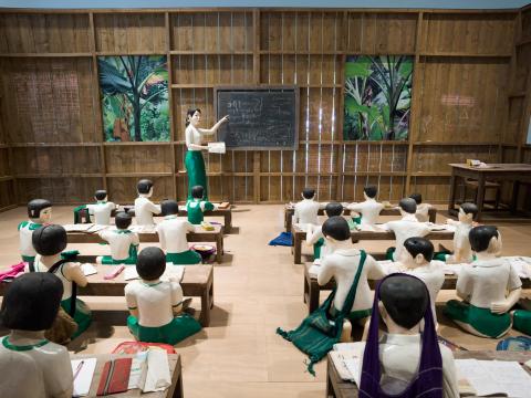 A close-up installation view of a sculptural work depicting a Myanmar school classroom with children seated at low benches, and with a teacher at a blackboard.