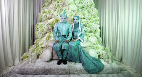 A photograph of a wedded couple wearing teal green outfits, including knitted teal balaclavas covering their faces. They sit on a light green throne-like seat with an enormous green floral arrangement and curtains behind them.