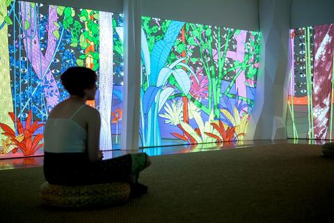 An installation view of a visitor sitting cross-legged in a gallery space that has brightly coloured animated forest scenes projected onto the walls.